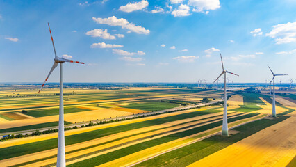 Wind turbines spin in a large green field under a blue sky with clouds. Wind turbines are turning in a vast field with crops during the day. The scene shows farmland and clear skies above. © Daniil