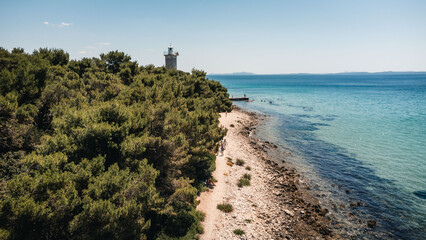 Obraz premium Coastal lighthouse on pine covered cliff above rocky shoreline and turquoise sea, seen from high aerial view with clear blue sky, bright sunlight, and tranquil Mediterranean landscape