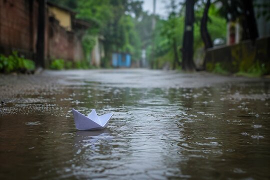 A small white paper boat floating on a rain-filled puddle along a quiet village road, gentle raindrops creating ripples, soft overcast light