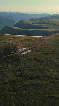 Vertical Screen: Beartooth Highway cuts through vast Montana USA alpine terrain with snow patches dotting green meadows and layered mountain ridges fading into hazy horizon at golden hour.