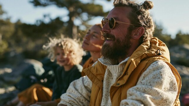Man with a beard and sunglasses sitting on a rocky hillside. he is wearing a yellow jacket and has a white sweater underneath.
