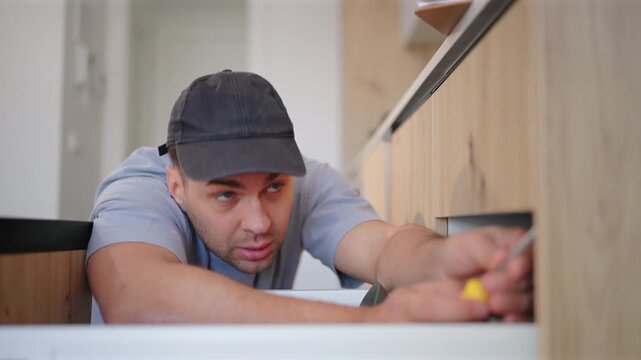 Maintenance professional with toolbox works in kitchen of home for repair tasks. Man wearing cap and gray shirt focuses on fixing drawer mechanism in modern wooden cabinetry interior.