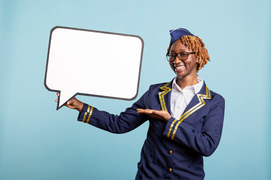 Smiling flight attendant stands against blue background, holding blank signboard. Confident air hostess gesturing to white speech bubble for promoting travel deals and airline marketing content.