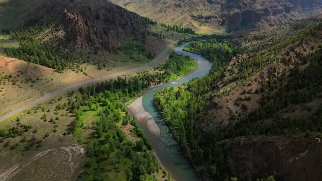 Aerial Montana USA view of turquoise river winding through dramatic canyon with rugged cliffs and pine forests. Scenic highway curves along valley floor beneath towering rock formations in summer.