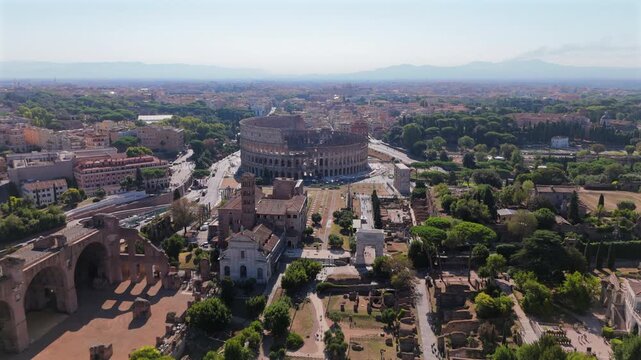 Cinematic aerial view in Rome Italy showcasing the iconic Colosseum amphitheater and Roman Forum ruins. Drone footage captures ancient architecture amid lush green trees and historic cityscape.