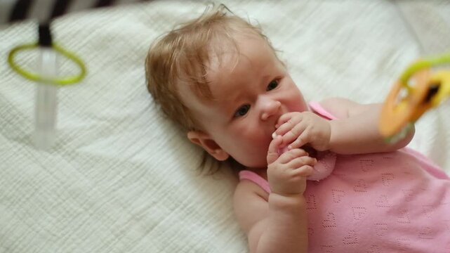 Cute baby girl in pink bodysuit lying on soft blanket and chewing silicone teether, authentic lifestyle portrait of infant teething at home.