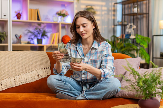 Young woman on home sofa counts dollar cash with happy smile and feels relief after income and success. Girl on home sofa smiles with savings pride after paid work for future