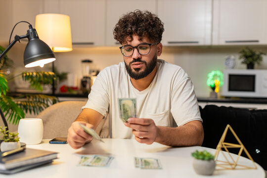 Arabian man at home counts dollar money cash uses calculator checks bills marks paid tracks saving. Egyptian guy at table sorts piles for spend and save plans budget adds note for month late extra.