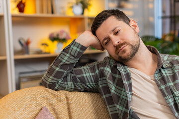 Obraz premium Young man on home sofa falls asleep from exhaustion with closed eyes and tired pose after stress routine. Adult guy on home sofa drifts into rest in living room while coping with fatigue and burnout.
