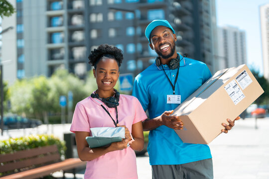 Two delivery workers stand outside in a city area. They hold packages, wear headsets and looking at camera. They look happy and ready to continue their work in the sunny weather.