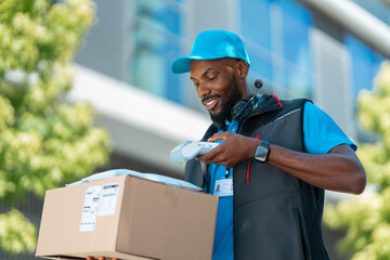 A delivery worker stands outside a modern building. He holds a box and uses a scanner to check the package. Trees are visible in the background on a clear day. © Videophilia