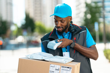 A delivery worker stands outdoors in a city setting. He scans packages while holding a handheld device. The scene shows tall buildings and people in the background. © Videophilia
