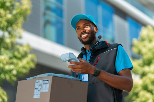 A delivery worker stands outside a modern building. He holds a box in one hand and a scanner in the other. He looks happy while checking the delivery information.