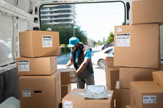 A delivery worker stands inside a van filled with brown boxes. He organizes the packages while parked in a city area with tall buildings around. It is daytime and sunny.