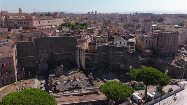 Aerial drone footage captures the ancient Forum of Augustus ruins in Rome Italy on a sunny day with clear blue skies, revealing weathered stone walls and columns amid vibrant historic buildings.