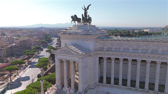 Aerial drone shot in Rome at Piazza Venezia featuring majestic Altare della Patria monument with bronze quadriga statue. Roman Forum ruins and Colosseum visible in the historic cityscape background.
