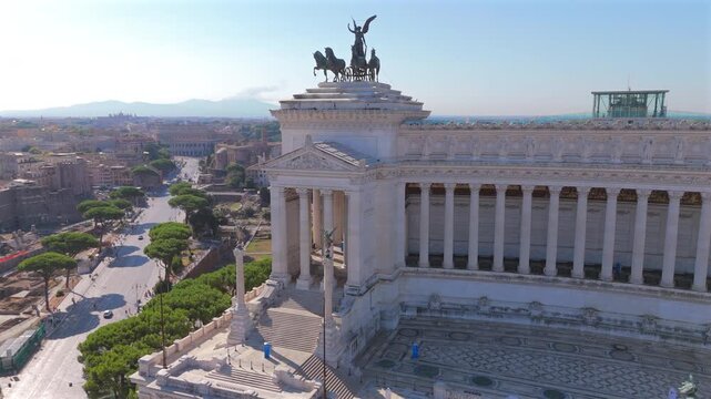 Aerial drone shot in Rome Piazza Venezia showing majestic Altare della Patria monument with Corinthian columns and bronze quadriga statues. Roman Forum ruins and Colosseum visible in the background.