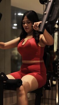 A woman in a red sports bra and shorts performs a chest press exercise on a gym machine, focusing on her workout.