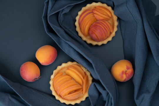A top view of tartlets shaped like small apricot cakes. Ripe apricots lie next to them. A cloth is draped around the tartlets.