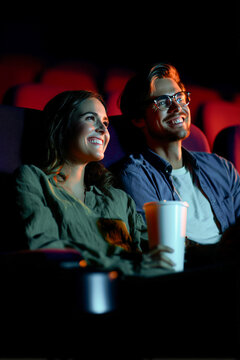 Happy young couple watching a movie in a dark cinema hall with a drink cup in hand. Smiling man and woman enjoying film premiere during a romantic date night