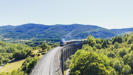 Old passenger train creating smoke while crossing historic arched stone viaduct through a green mountain landscape on a sunny day © DMegias