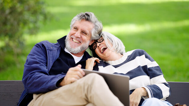 Happy senior couple laughing while sitting on a park bench, looking at a device and enjoying togetherness in nature