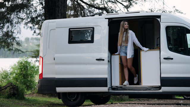 Woman standing in doorway of a white camper van, looking out at the calm lake and surrounding forest
