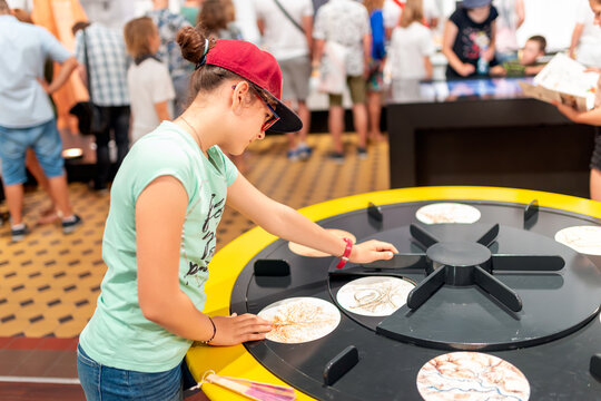 Side View of a Focused Young Girl in a Red Cap Interacting with a Round Educational Exhibit Wheel Inside a Busy Science Museum