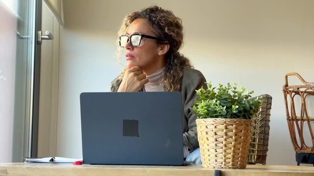 A focused woman with curly hair and glasses works diligently from her home office, taking notes and typing on her laptop, embodying modern productivity and remote work.