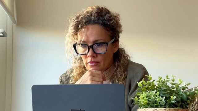 A focused woman with curly hair and glasses works on her laptop, then pauses for a thoughtful moment, looking out the window, reflecting on her work. This scene captures remote work and contemplation.