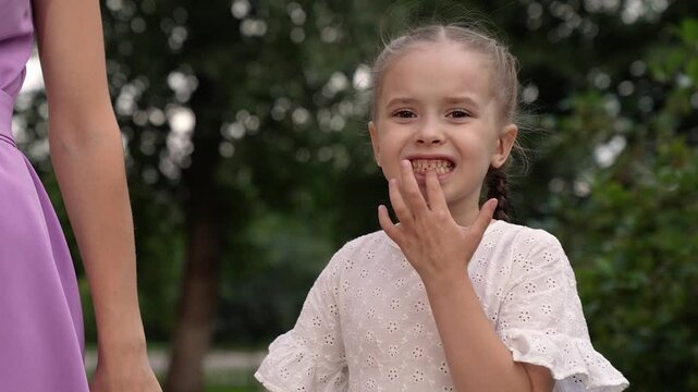 Happy little daughter shows gap on milk teeth place holding mother hand in green park. Cute daughter without lower teeth holds mother hand during walk in park. Family of mother and daughter in park