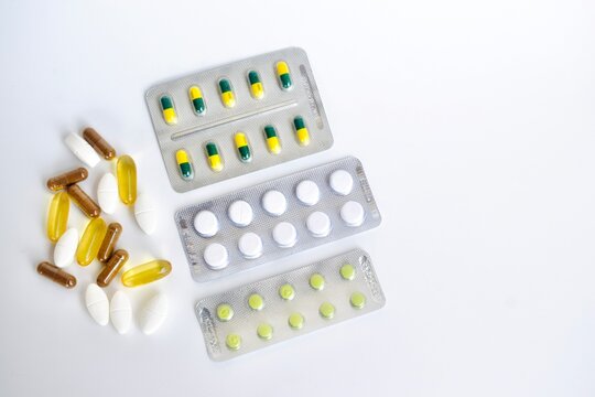 Top view showing a collection of white tablets, yellow softgels, and brown capsules scattered adjacent to medical blister sheets on a white seamless backdrop.