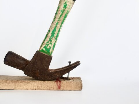 Close-up studio shot of a rustic claw hammer with a green weathered handle pulling a metal nail out of a wooden block against a white background.