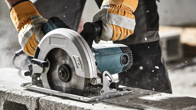A construction worker wearing protective gloves uses a circular saw to cut a heavy concrete block.