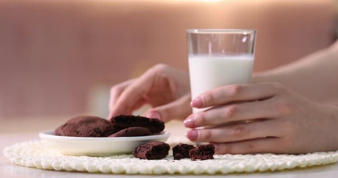 Woman putting glass of fresh milk onto table and taking tasty chocolate cookie from plate in kitchen, closeup. Camera moving