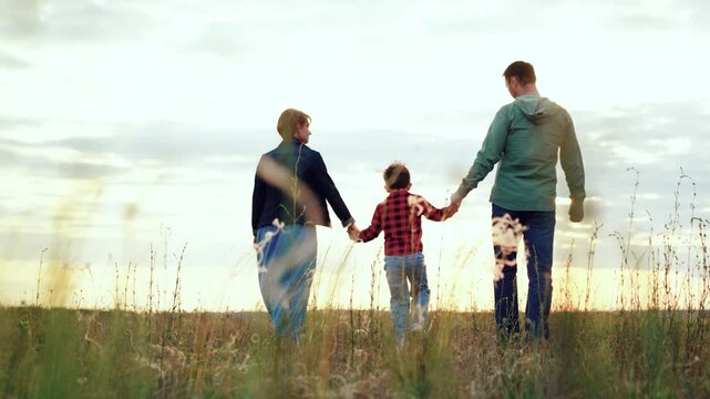Small family enjoys leisurely walk in field basking in joyous moments brought by windy weather on holiday. Family hold hands walking across grassy valley. Family vacation and active pastime
