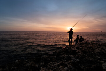 Fototapeta premium Silhouette of Fishermen Fishing on Rocky Coast at Sunset with Dramatic Ocean View, Sunset Fishing on Rocky Shore with Silhouette Anglers