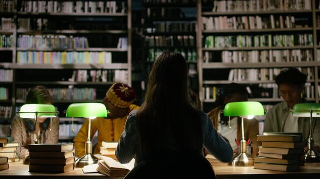 Back view of young woman with headphones seated at quiet library table with open books, green lamps and classmates studying in background, late night study session, for education technology.