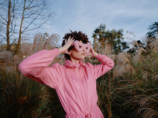 Fototapeta premium Portrait of a person in a pink outfit adjusting a playful mask in tall grass outdoors under a clear sky, showcasing creative fashion and bold nature photography with a carefree stance against a
