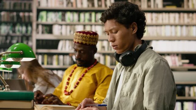 Man student with headphones reads open book in library study area, framed by green lamps and books in quiet academic environment, study session.