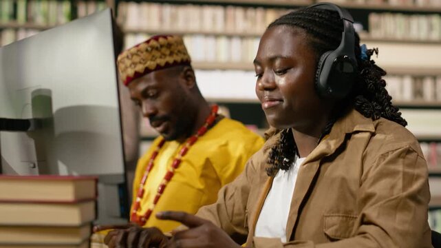 Smiling black student wearing headphones and typing at desktop monitor with books and classmates nearby in warm library studying space for coursework.