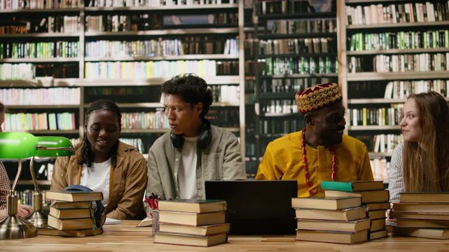 Diverse students discuss classwork during group study around library table with open books, textbooks and green lamps, collaborative academic study, peers collaboration, for online learning.