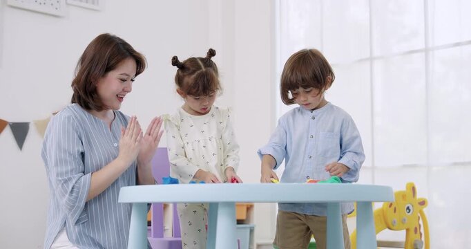 Asian caregiver helping preschool boy, preschool girl press modeling clay using cutter tool on round table near window light, creative play lesson, sensory activity, kindergarten learning moment