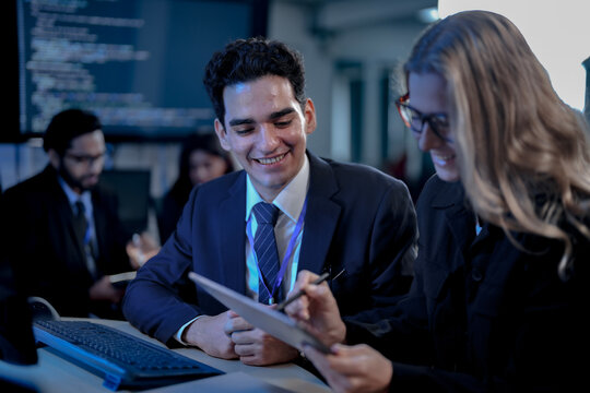 Cheerful young developer showing tablet results to his smiling blonde colleague comparing release notes seated close together in a dark blue artificial intelligence startup office together.