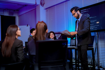 Obraz premium Executive standing at a desk reviewing documents with his team confirming the submission time in a dim blue technology office late at night captured in an authentic working moment.