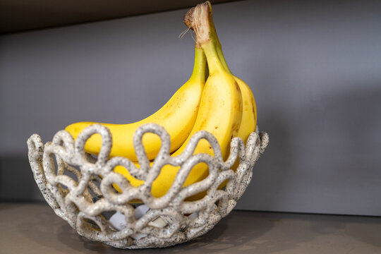 Ripe yellow banana bunch resting in a decorative white coral shaped ceramic bowl against a neutral wall on a kitchen shelf