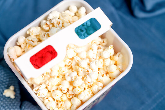 Close view of fresh buttery popcorn in a white tub with red and blue 3D cinema glasses on top ready for a movie night snack