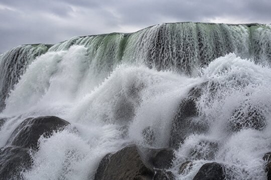 Waterfall top, massive water flow crashing onto rocks, white spray, overcast daylight.