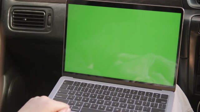Person typing on laptop with green screen inside car. Hands visible, focused on keyboard. Green screen perfect for chroma key editing. Ideal for travel, work from car, or mobile office scenes