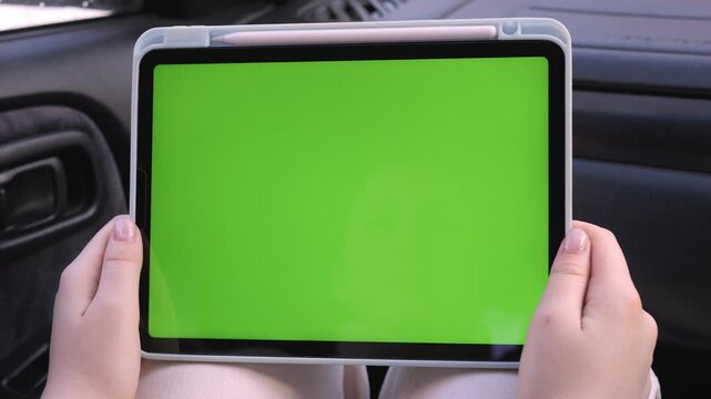 Hands hold tablet with green screen inside car. Person focuses intently on screen content. Useful for app demos, digital marketing, or travel ads. Perfect for overlaying custom graphics or text
